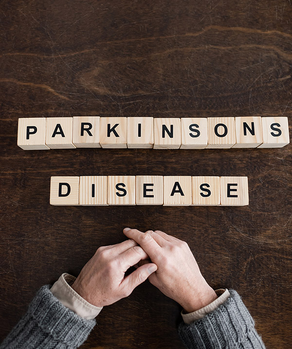 Wooden blocks spelling "PARKINSONS DISEASE" on a dark wooden table with clasped hands below.