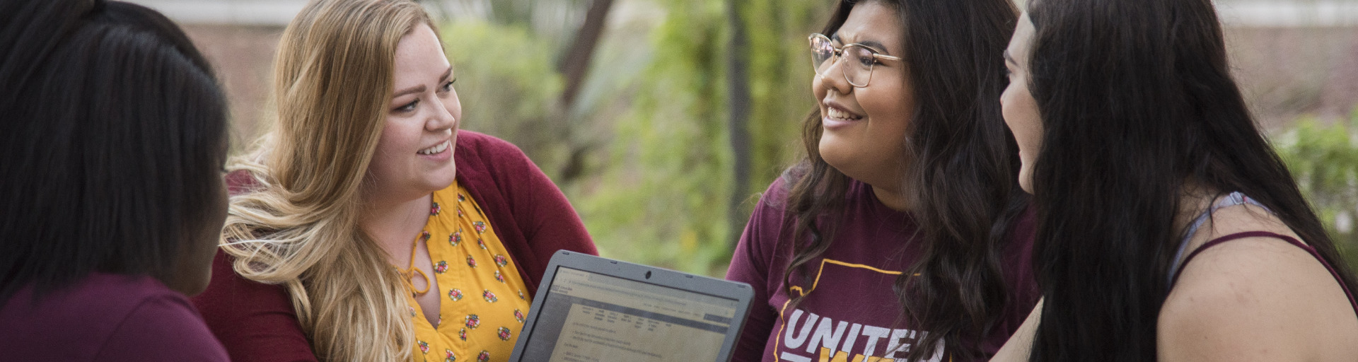 Four students sitting together outdoors, smiling and talking around a laptop.